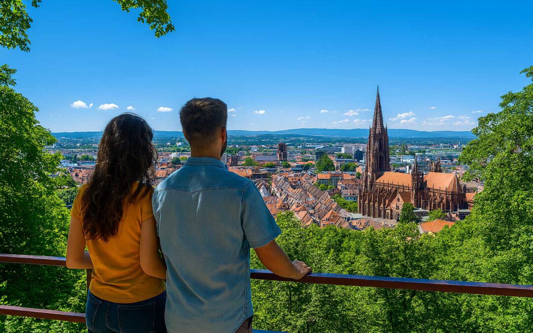 Ein Mann und eine Frau stehen an einem Aussichtspunkt über Freiburg und blicken auf die Altstadt mit dem Freiburger Münster. Sonniger Himmel, grüne Hügel und rote Dächer prägen das Panorama.